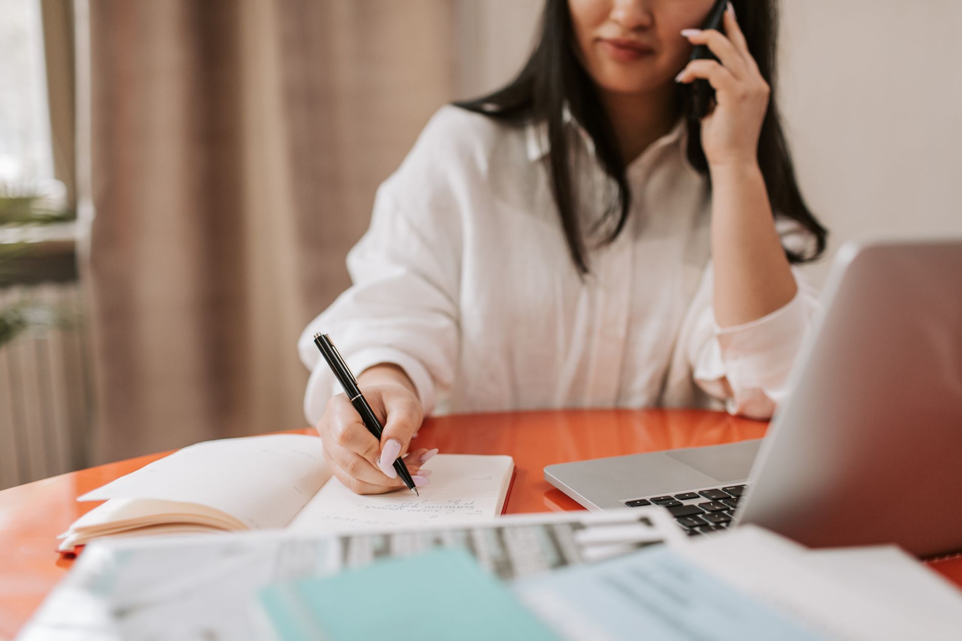 femme assise à son bureau, au téléphone, qui prend des notes sur un carnet devant un ordinateur ouvert