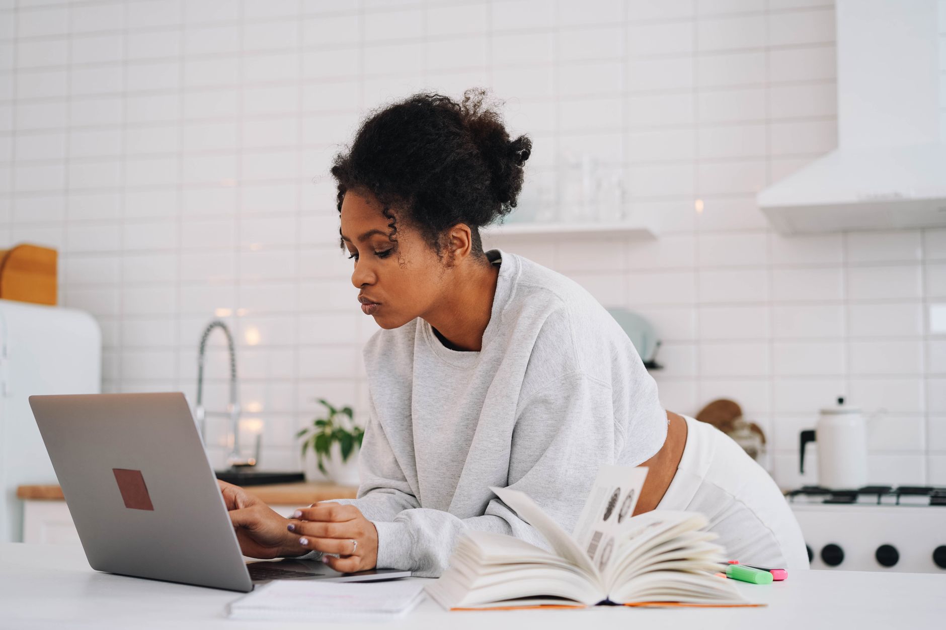 femme assise à la table de sa cuisine, devant un ordinateur portable et des livres ouverts, qui prépare son dossier de reprise d'études
